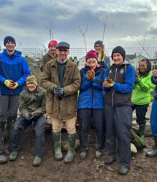 A group of volunteers stand on muddy ground to pose for a photograph, each holding a tree sapling aloft.