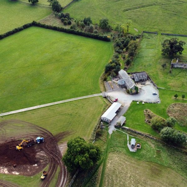 Aerial view of a large circular scrape being excavated by digger on green pastureland.