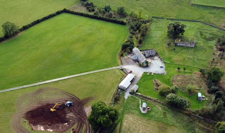 Aerial view of a large circular scrape being excavated by digger on green pastureland.