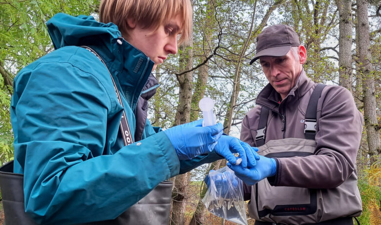 Two men are in outdoors clothes and waders. They are wearing blue latex gloves. One has a syringe in his hand and the other a clear plastic bag containing water and a vial.