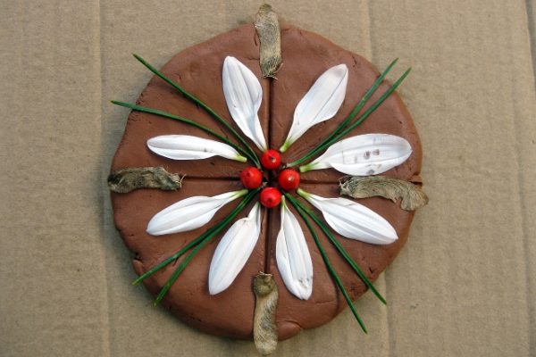 A piece of artwork created with a flat stone with while petals, red berries and green fir needles arranged on it in the shape of a flower.