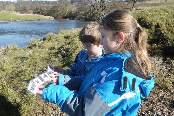 Two young people standing by a river bank looking at a guide telling them about the flora and fauna they may see around the river.