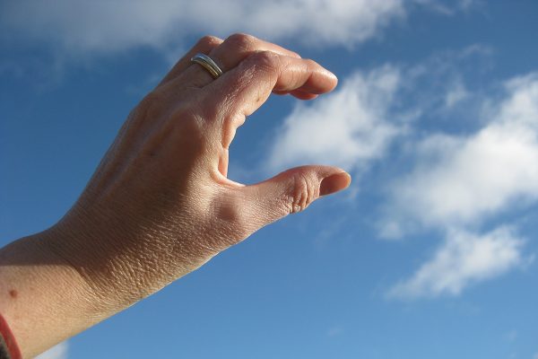 Close up of a hand being held up to a blue sky pretending to catch a wispy cloud between thumb and index finger.