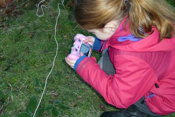 A young person in pink coat bends down, holding a pink camera to take a picture of grassy ground.