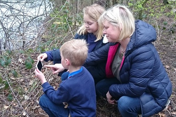 Two young people and adult crouched by the edge of a water course and sparse foliage at the water's edge are examining an image captured on a mobile phone.