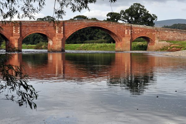 View looking from river bank, along its course to a stone arched bridge crossing the watercourse.