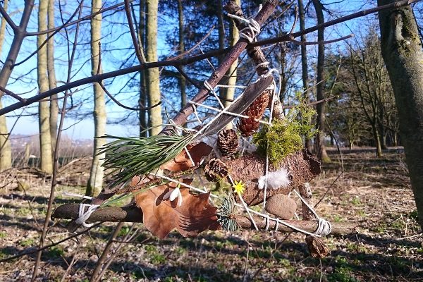 Small sculpture created from a triangle of sticks tied with string and adorned with autumn leaves, mosses, fir needles and small flowers, sited in an autumnal woodland setting.
