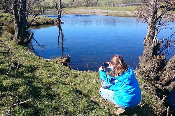 A young person crouched by the edge of a water course on a sunny autumn day, pointing a camera to take a picture.