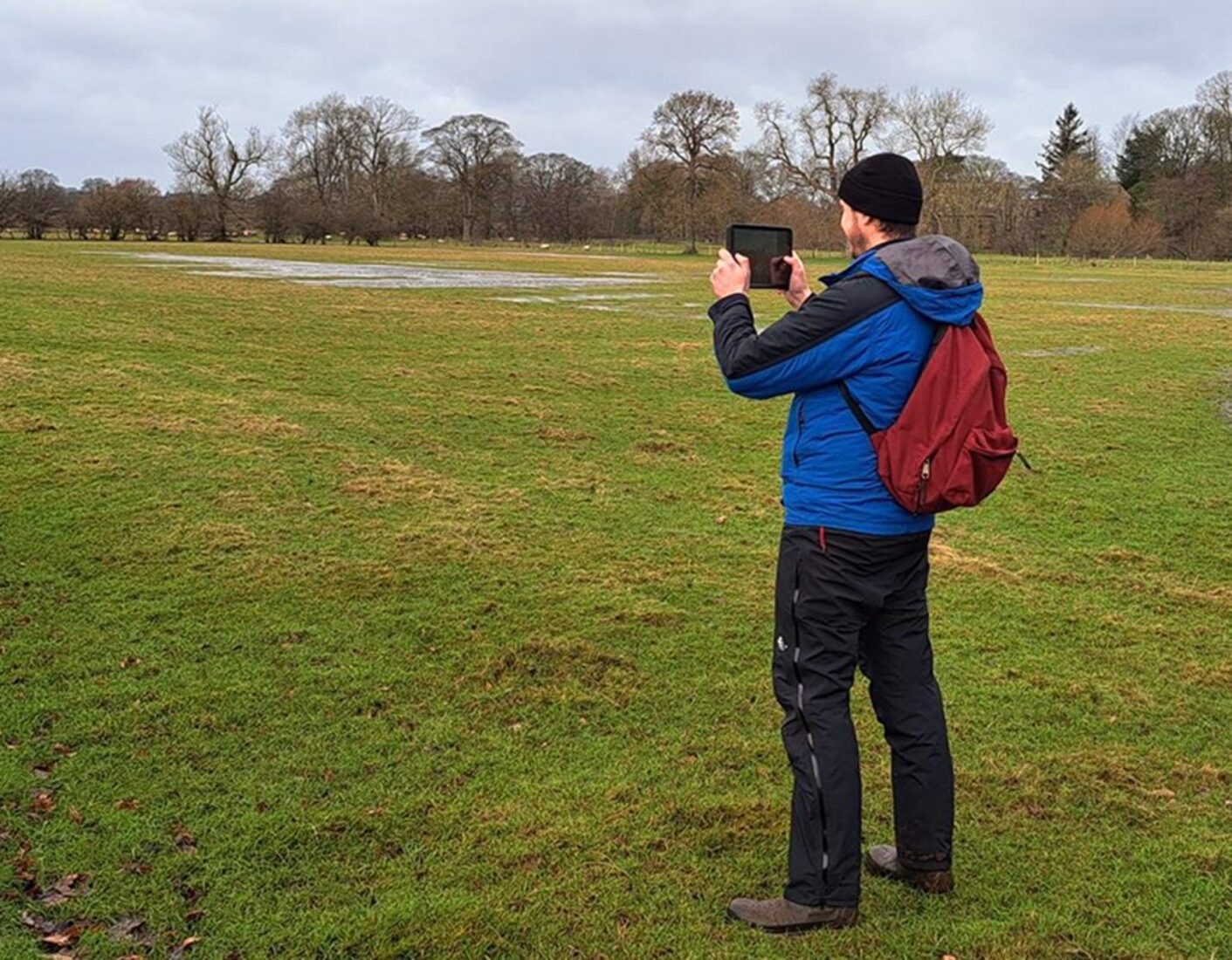 Member of staff in field holding up an electronic tablet and taking a picture of the scene.
