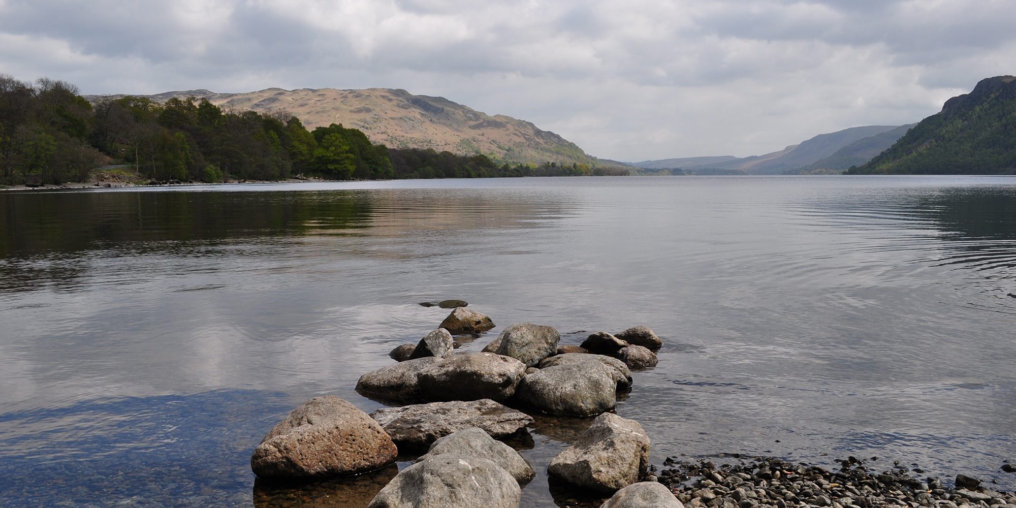 It's a calm day on Ullswater lake - can see the fells and forests in the background reflected in the water. In the foreground are some stones leading out into the water.