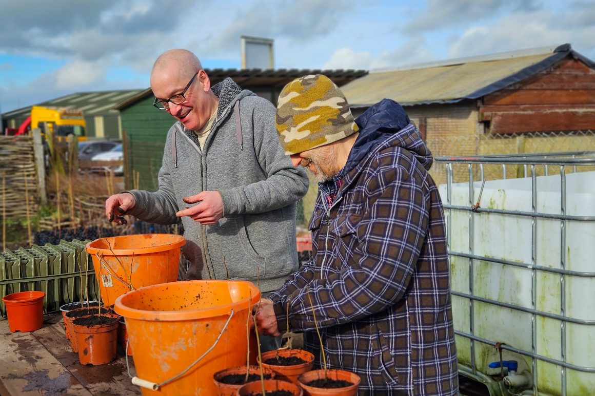 Two ERT volunteers stood at a wooden table, repotting saplings into larger pots using soil stored in large orange buckets. Onsite at the Penrith community tree nursery.