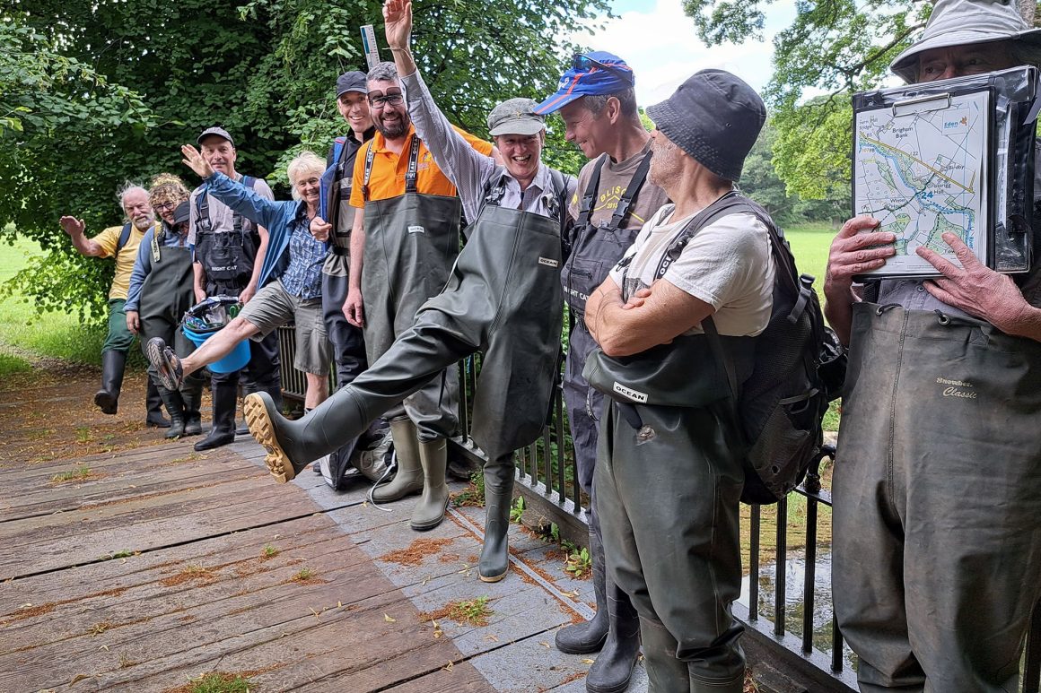 A group of people are standing against a metal barrier on a bridge. They are wearing waders. One woman is raising her leg and hand at a jaunty angle whilst another man hides behind his map.