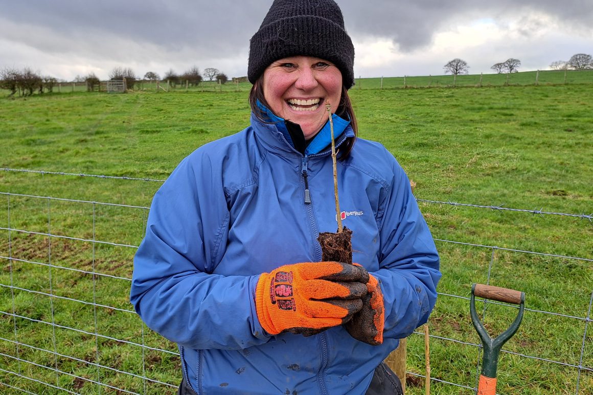 A woman holds a tiny tree sapling in her gloved hands and smiles at the camera. In a minute, she will plant it in the ground.