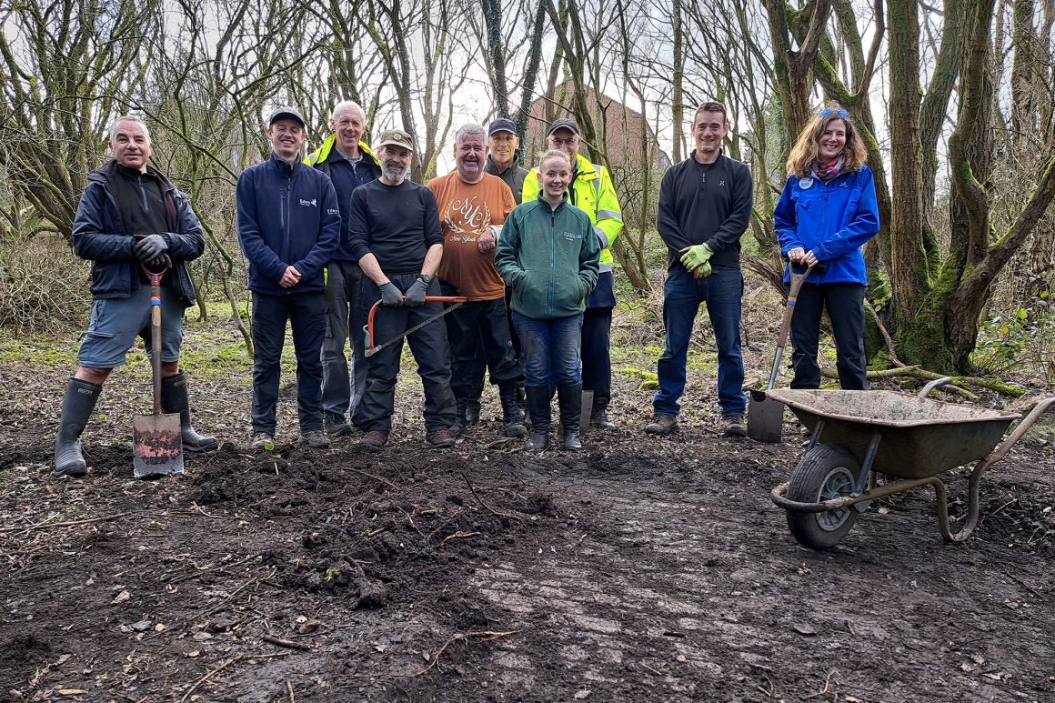 A group of people are stood in a line. Two of them are holding spades. On the ground in front of them there are cobblestones showing where the people have dug away a layer of earth that was hiding them.