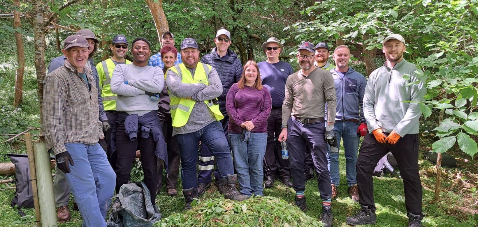 A group of volunteers posing for a photograph stood behind a pile of Himalayan balsam uprooted and piled up during a Balsam bashing event on the site amongst woodland adjacent to river.