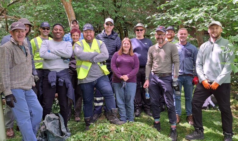 A group of volunteers posing for a photograph stood behind a pile of Himalayan balsam uprooted and piled up during a Balsam bashing event on the site amongst woodland adjacent to river.