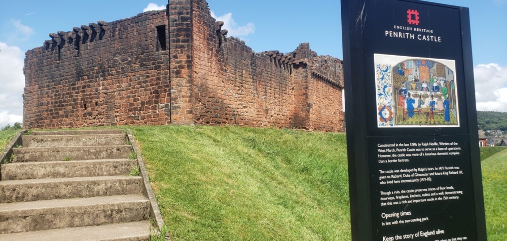 A view from foot of grass bank looking up at Penrith castle walls below a blue sky.