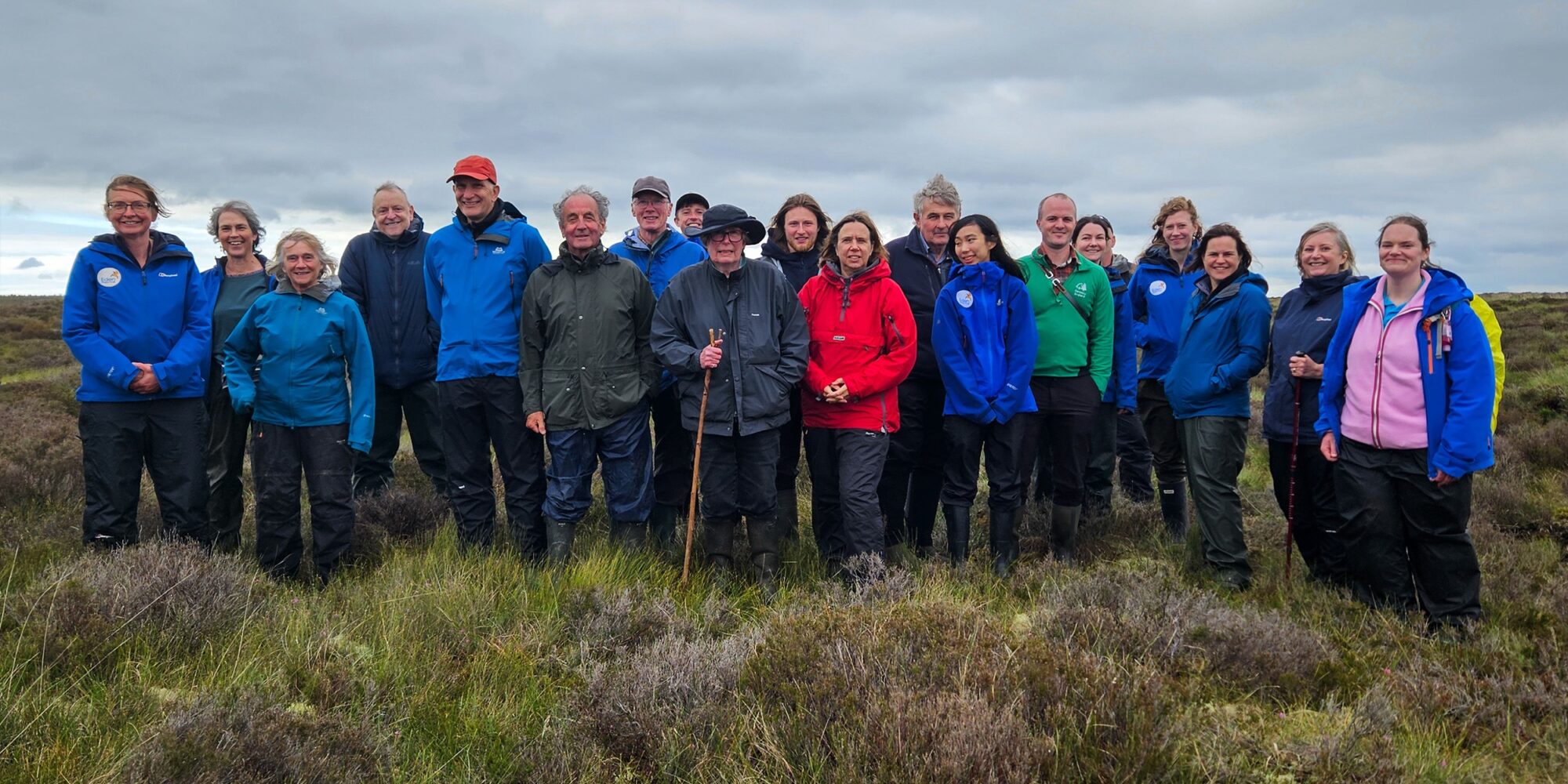 A large group of people are stood in two informal lines posing for the camera. It's a cloudy day and they are on the top of a moor