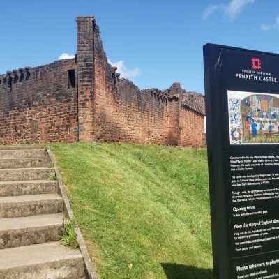 A view from foot of grass bank looking up at Penrith castle walls below a blue sky.