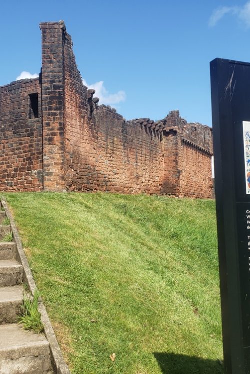 A view from foot of grass bank looking up at Penrith castle walls below a blue sky.