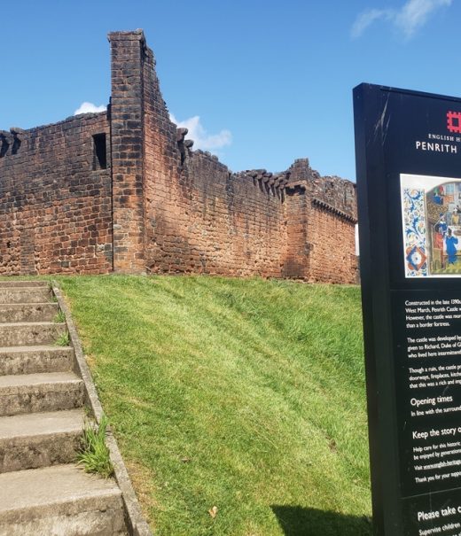 A view from foot of grass bank looking up at Penrith castle walls below a blue sky.