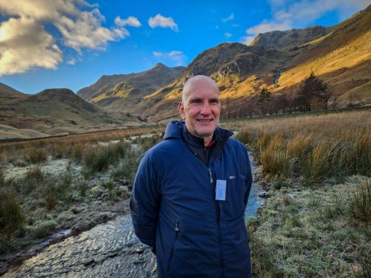 A man stands smiling at the camera. It is a cold, sunny winter's day and he is wearing a thick blue waterproof coat. Behind him are jagged fells bathed in sunshine.