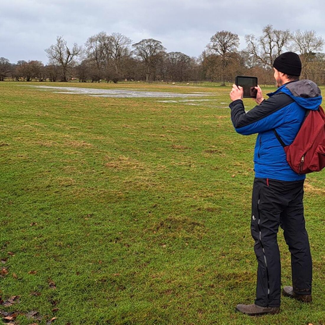 Man staring across grassy field holding a tablet up to take a photograph of the scene.
