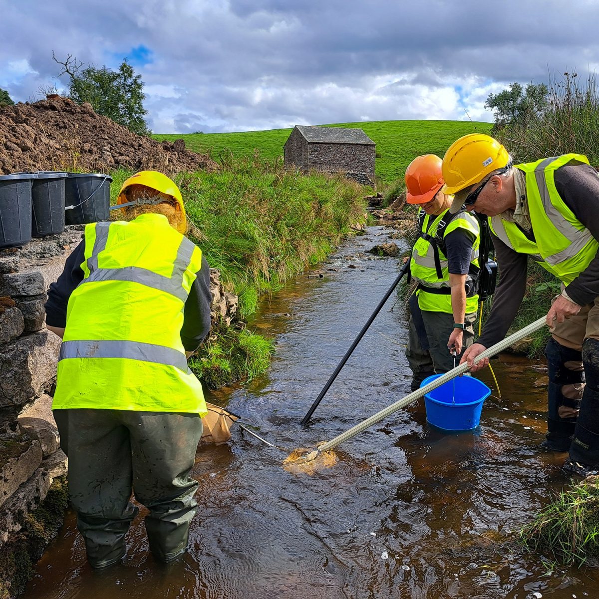 Three men in hi vis jackets and hard hats stood in shallow water channel with nets and buckets.