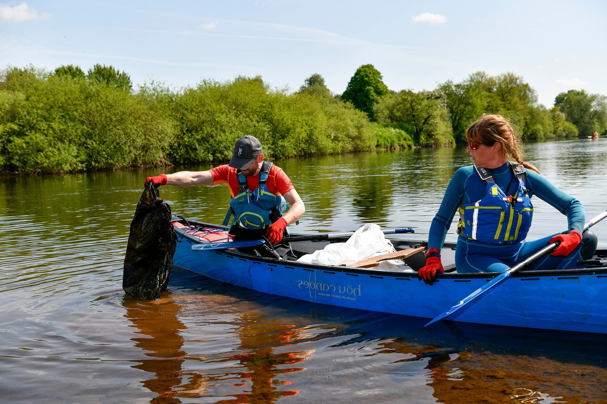 Two people in open canoe on a lake have stopped to fish out a large black bin bag from the water.