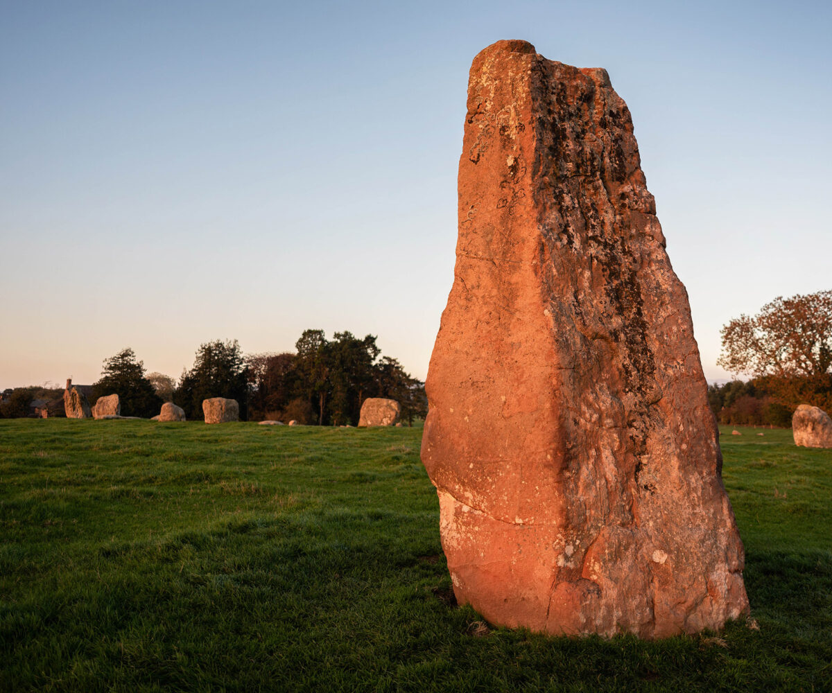 A large oblong stone stands outside of a circle of smaller stones; the red tones of the stone is brought out by the evening sun shining on it.