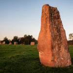 A large oblong stone stands outside of a circle of smaller stones; the red tones of the stone is brought out by the evening sun shining on it.