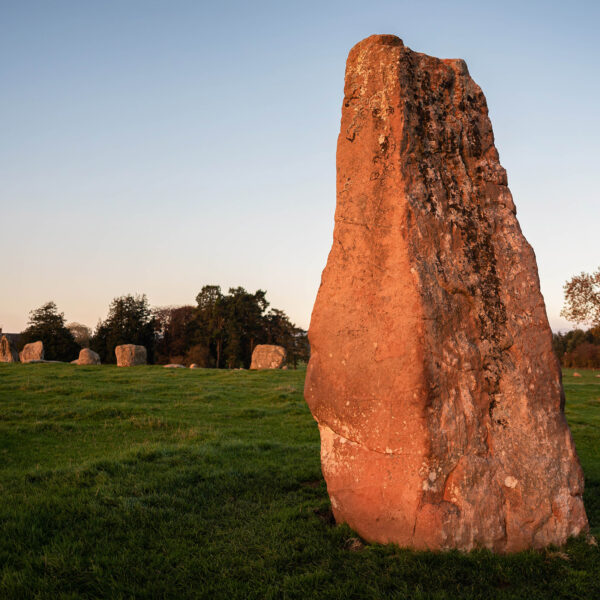 A large oblong stone stands outside of a circle of smaller stones; the red tones of the stone is brought out by the evening sun shining on it.