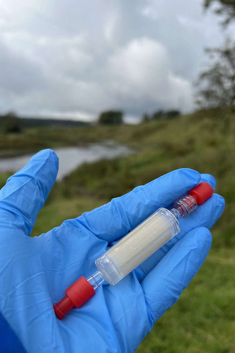 a tube with a red cap at both ends is being held by a hand wearing a blue glove. In the background can see a stream and grassy riverbank. It is a grey, cloudy day.