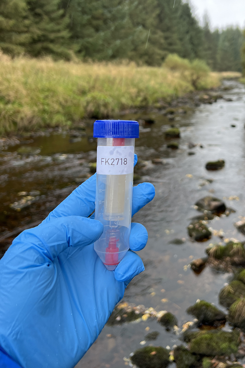 a hand wearing a blue glove is holding a tube that contains another tube with water in it. In the background there is a stream with rocks in it and a riverbank covered in grass leads down to it.