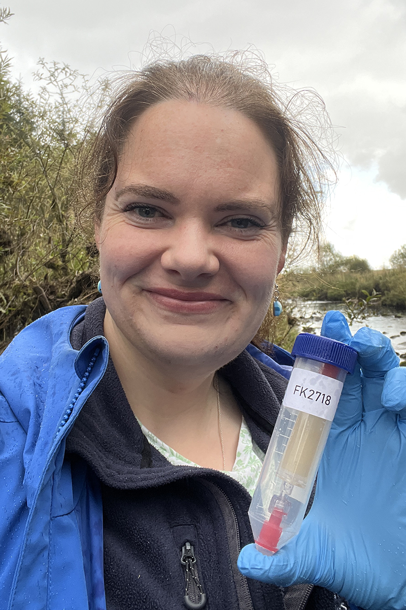 A woman wearing a blue coat holds a tube with a blue lid on it. Inside is another tube with water in it.