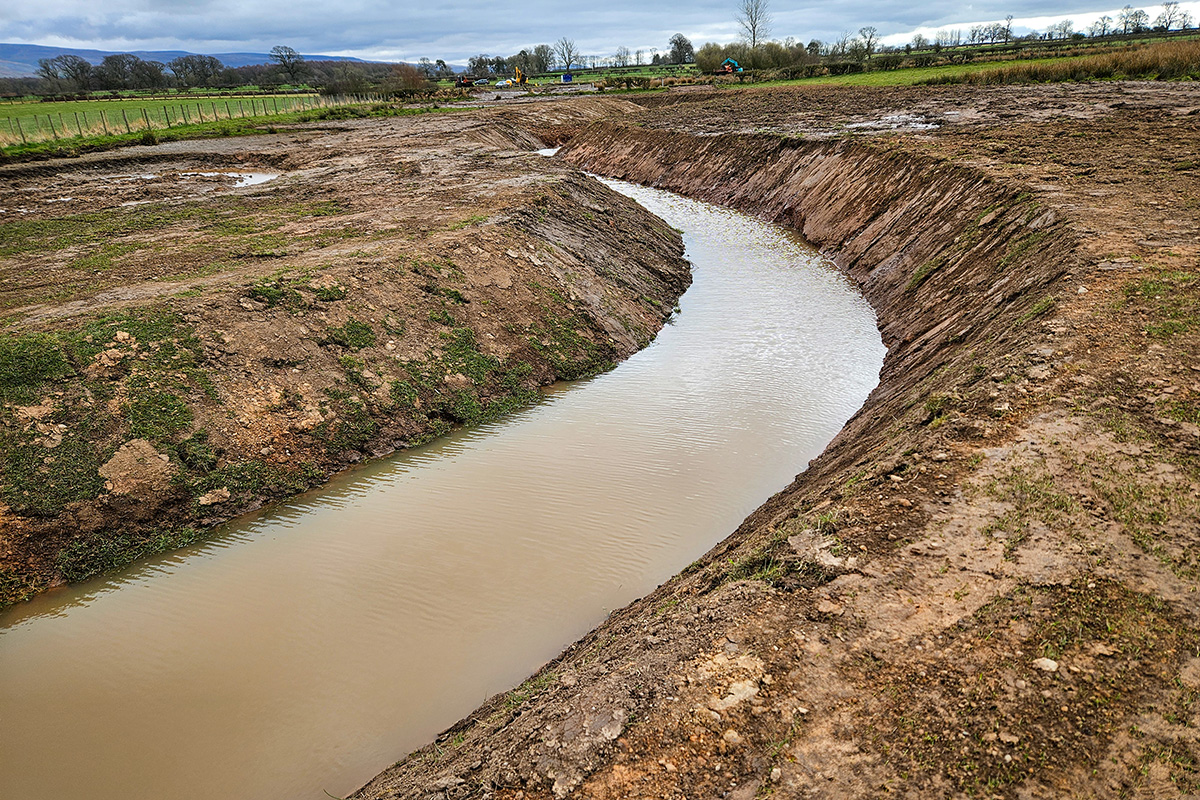 A curving river channel half filled with crown water. The banks are bare and can see the soil as the channel has recently been cut.